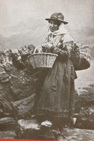 Copy of a postcard of a Llangwm Pembrokeshire fisherwoman in traditional costume standing on the River Cleddau fore shore holding a basket